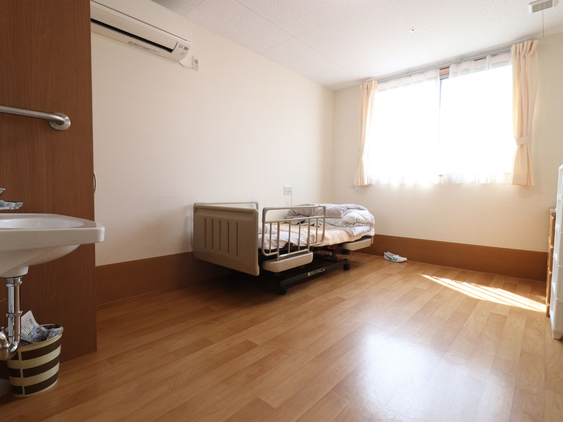腰高窓があるワンルームで、床はフローリングである。落下防止用の柵がついた電動ベッドで、壁際にある。入り口付近に洗面所が見える。