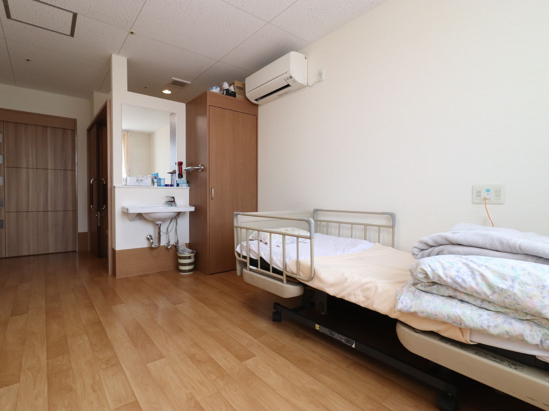 茶色の床、白い壁のワンルームには、洗面所、クローク、電動ベッドが置いてある。洗面所の棚には、歯磨き粉などが置いてある。