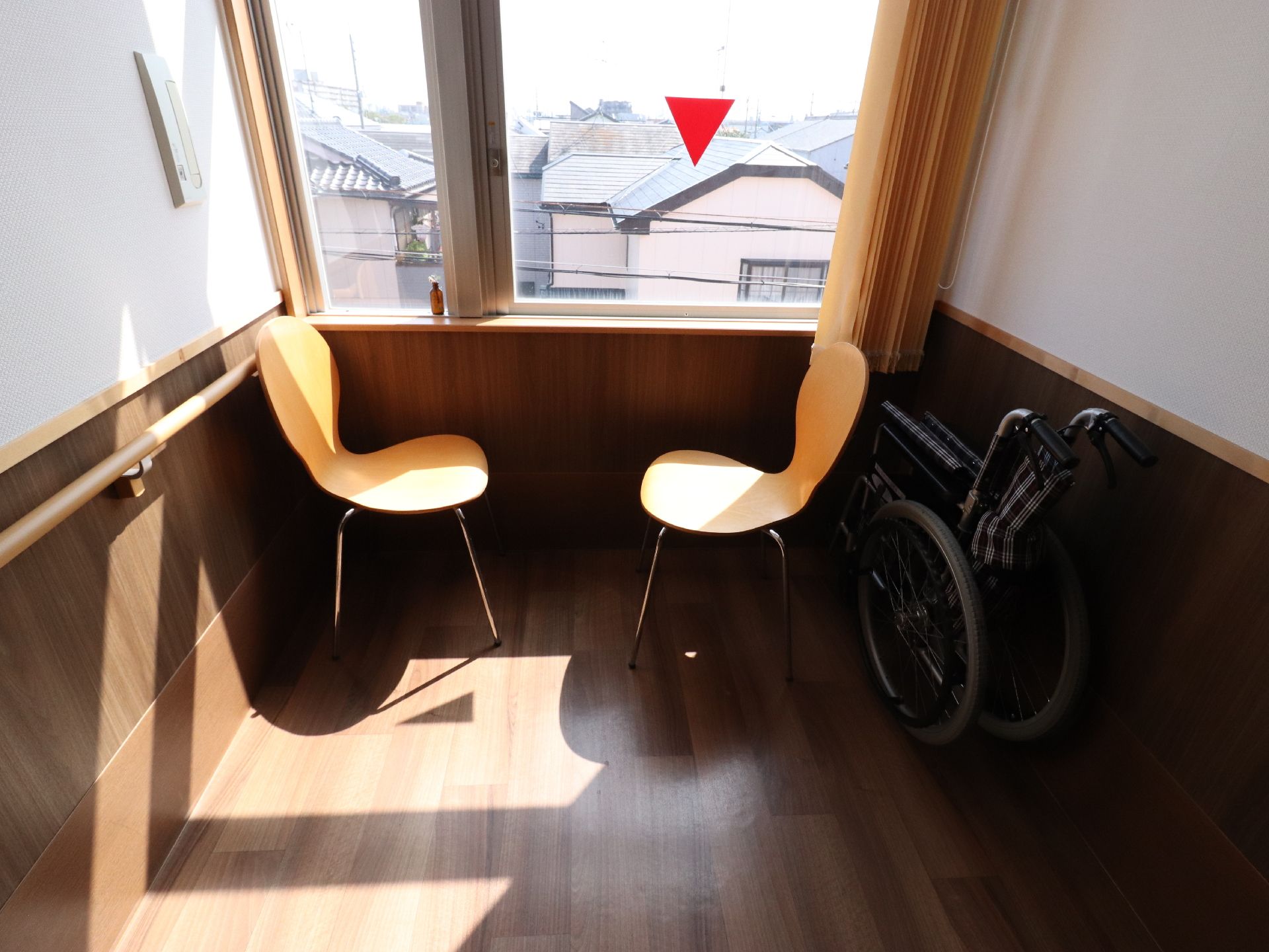 フローリングの床の上に椅子が2脚置いてあり、すぐそばに窓がある。家が建ち並んでいるのが見える。折り畳み式の車いすが置いてある。