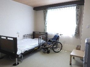 転落防止柵が取り付けられた介護ベッドの横に車椅子が置いてある。カーテンとレースカーテンが掛けられた腰高窓がある。