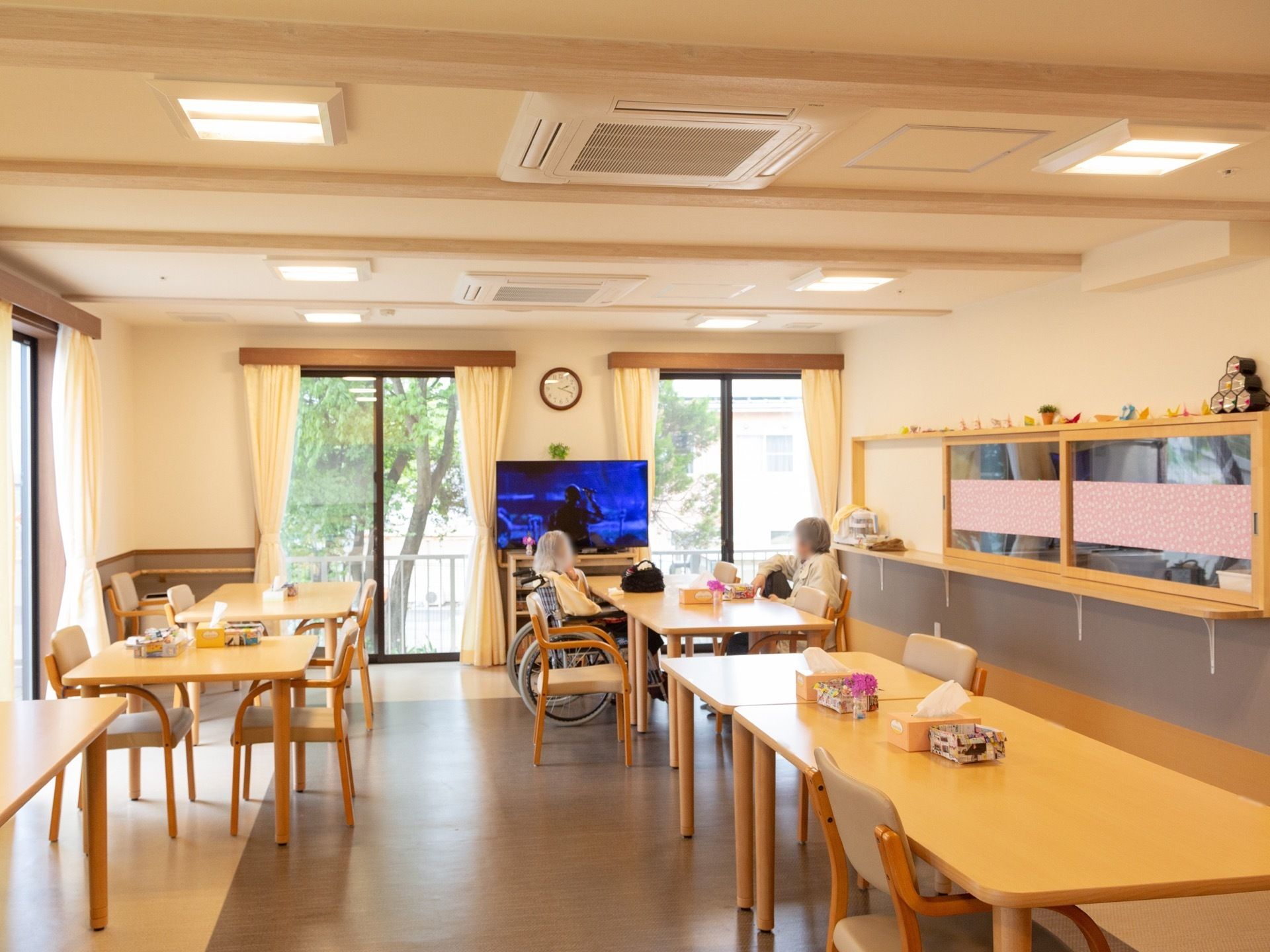 テーブルと椅子が整然と並べられ、入居者様がテレビを見ている。施設内の厨房でスタッフがおいしい食事を調理している。