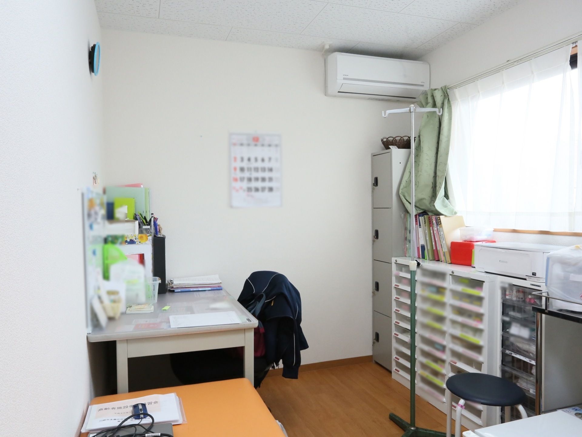 事務室風の室内にデスクや椅子、収納棚などが並んでいる。壁にはエアコンが設置されていて、カレンダーも貼られている。