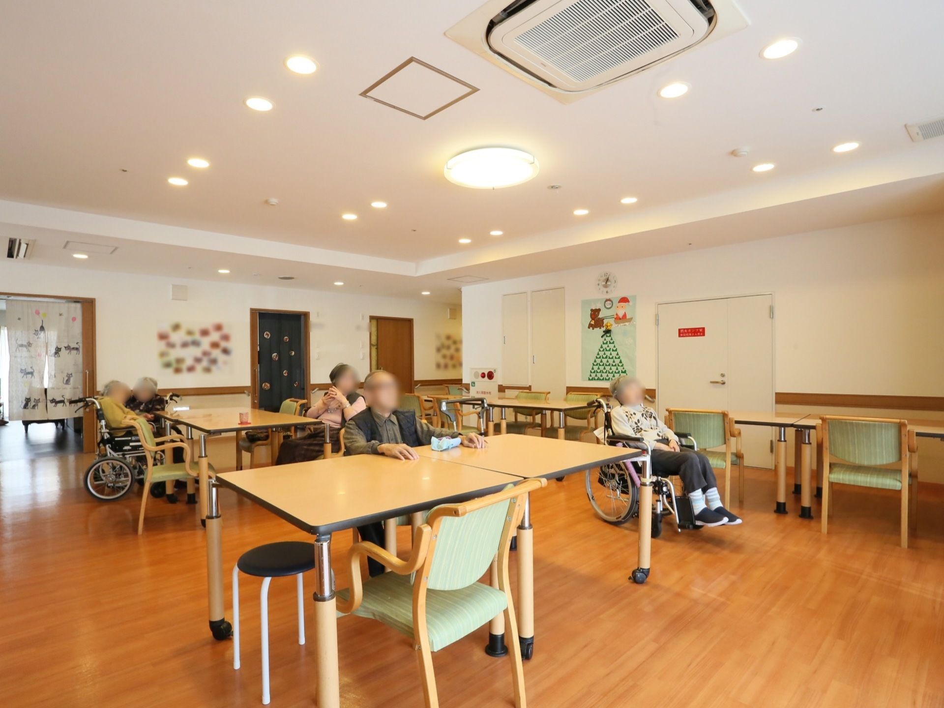 テーブルや椅子が多数置かれた共有スペース。入居者様が椅子に座ったり、車椅子に座ったりして、なごやかにテーブルを囲んでいる。