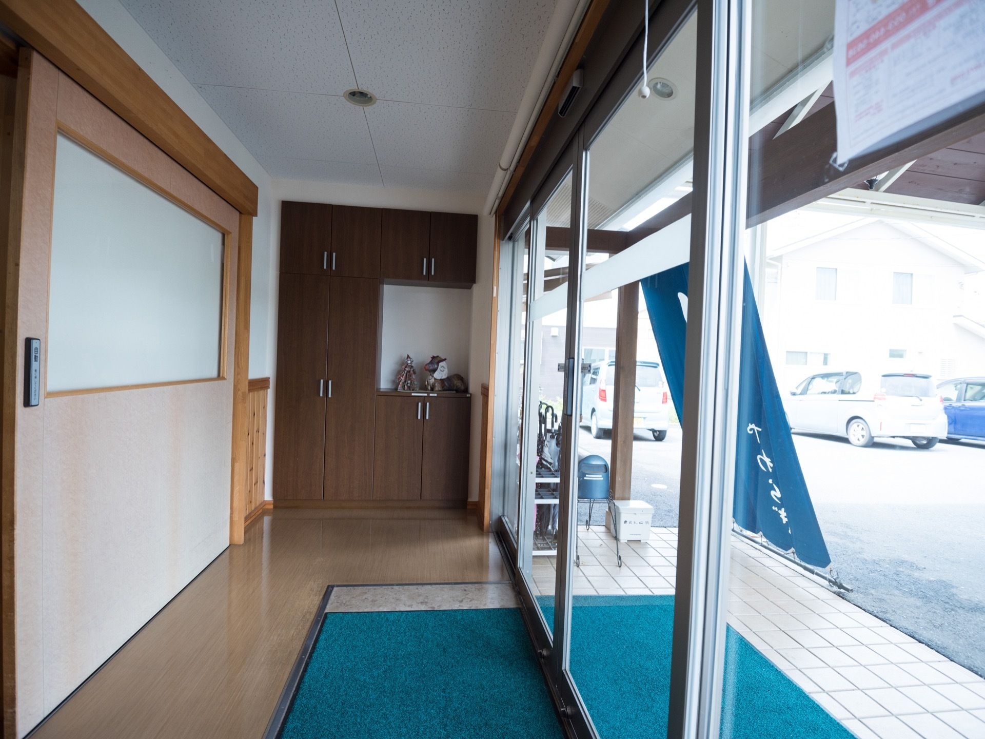 自動ドアの出入り口があり、床に緑のマットが敷いてあるエントランス。奥の壁にはこげ茶色の収納が設置されている。