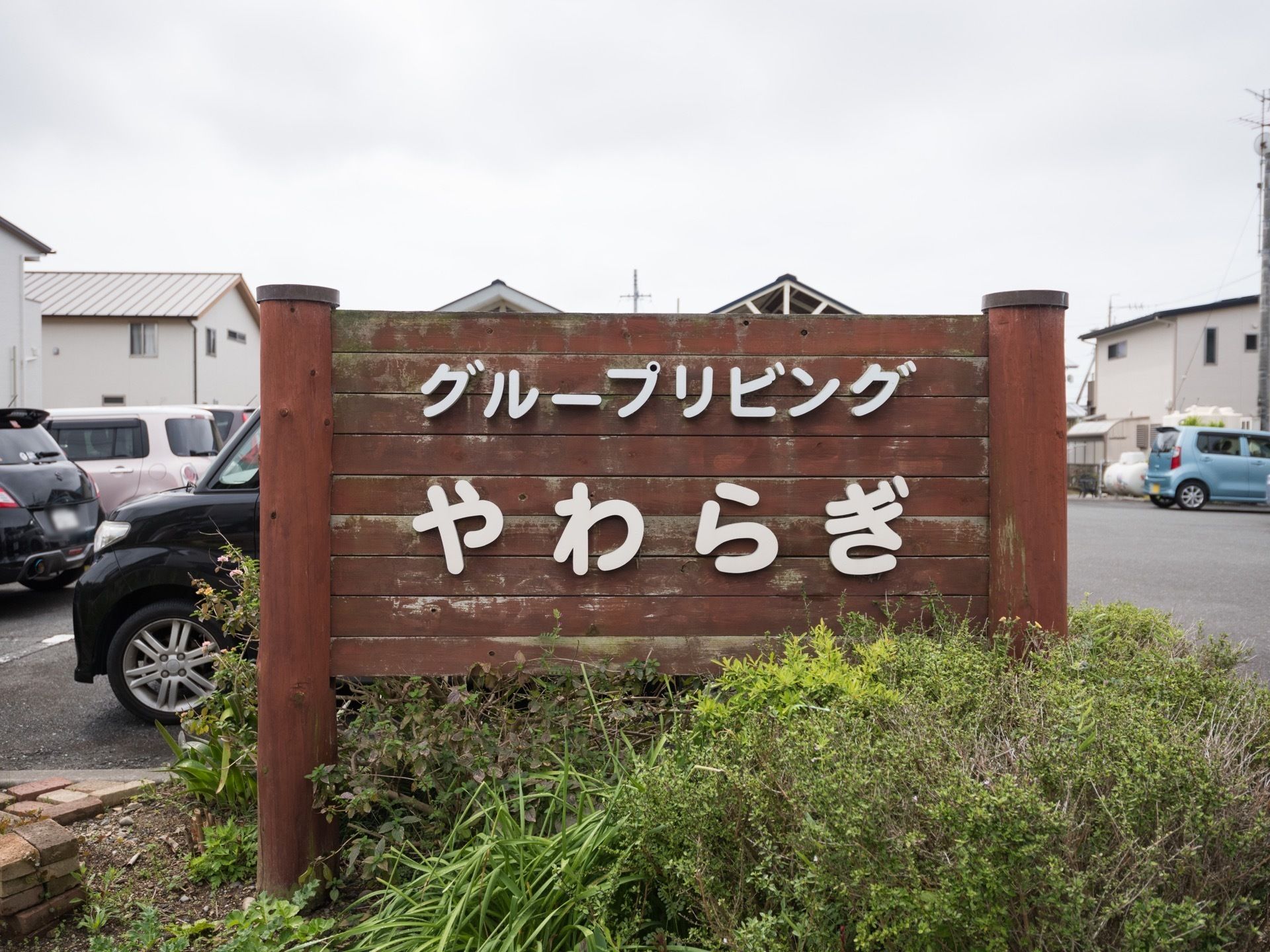 植え込みの中に茶色い木目調の看板が立っており、白い文字で施設名が書かれている。看板の後ろにあるスペースには車がたくさん停まっている。