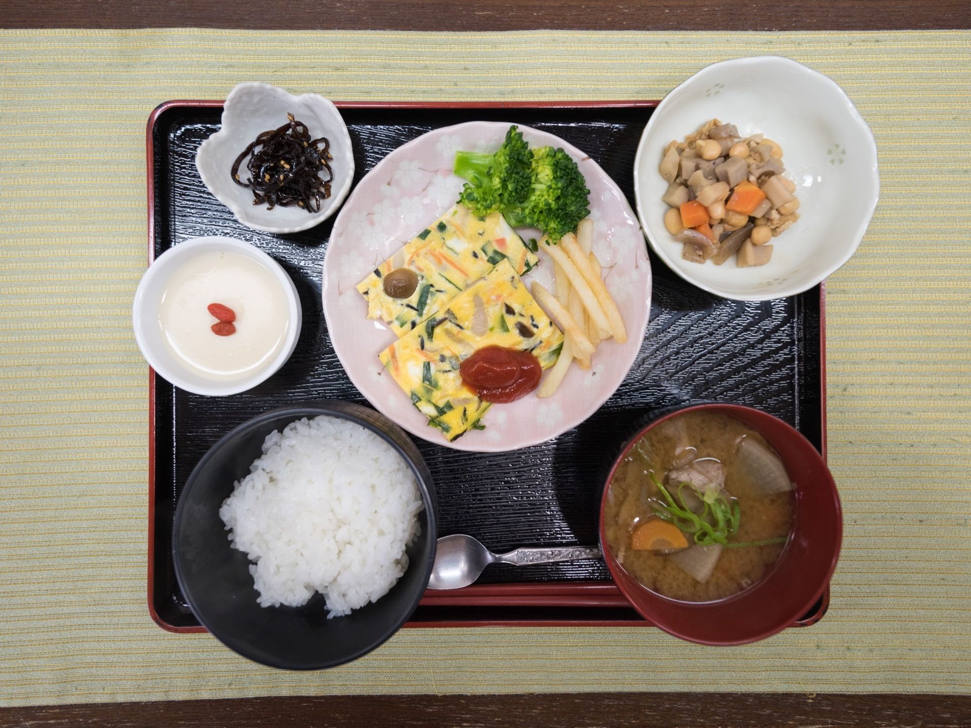 黒の四角いトレーに食事の用意がされている。ご飯や味噌汁、オムレツ、煮物などが並べられており、彩り豊な食事である。