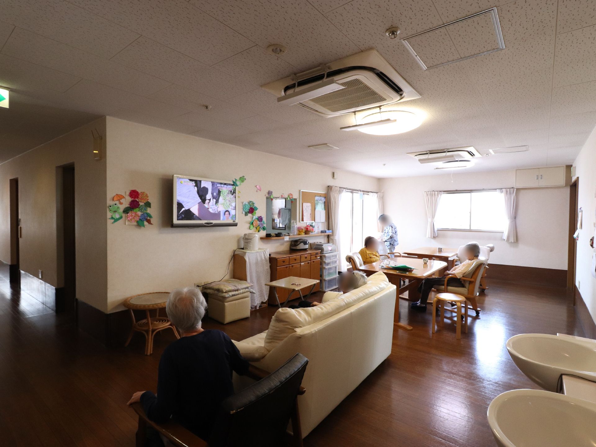 壁掛けテレビがある部屋には茶色いテーブルと椅子が数脚置かれている。その椅子と近くにあるソファーには入居者様が座っている。