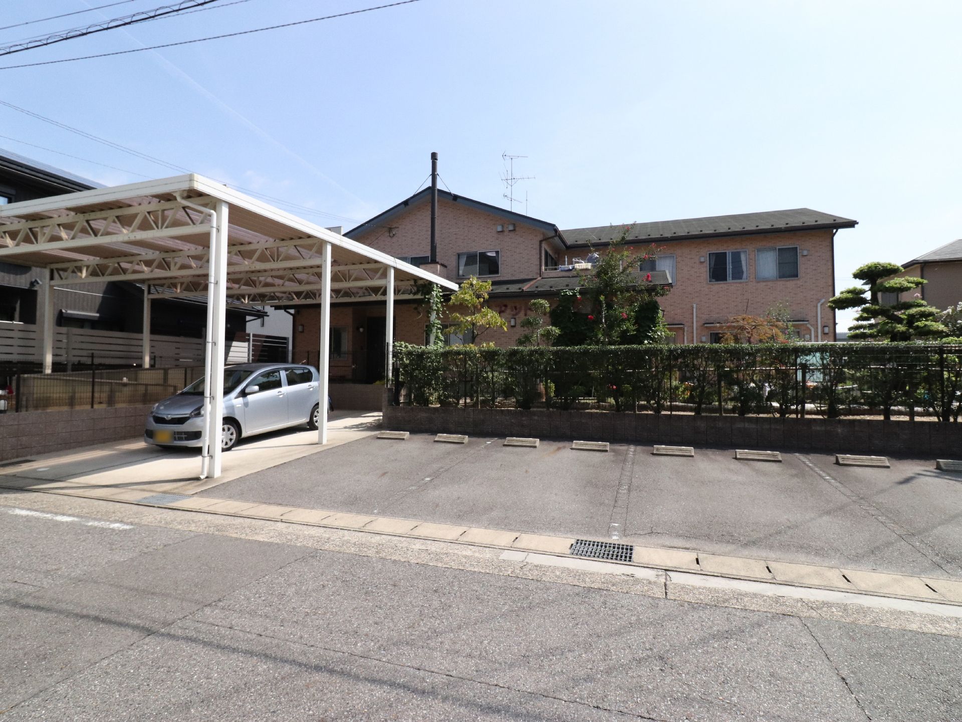 施設のすぐそばにはカーポートがある場所とない場所に分かれた車の駐車スペースがある。施設の周りは緑で囲まれている。
