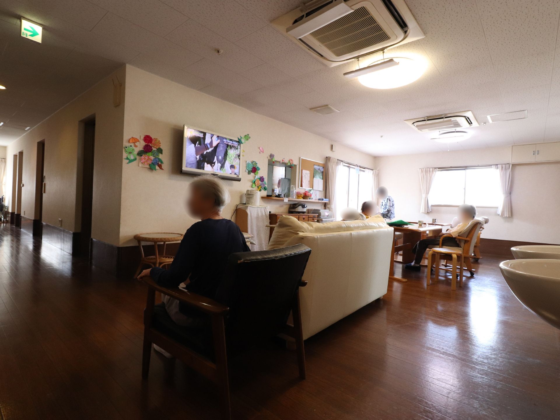 壁にはカエルと花の飾りが施されている。その横にはテレビがかけられている。周りには茶色いテーブルや椅子が置かれている。