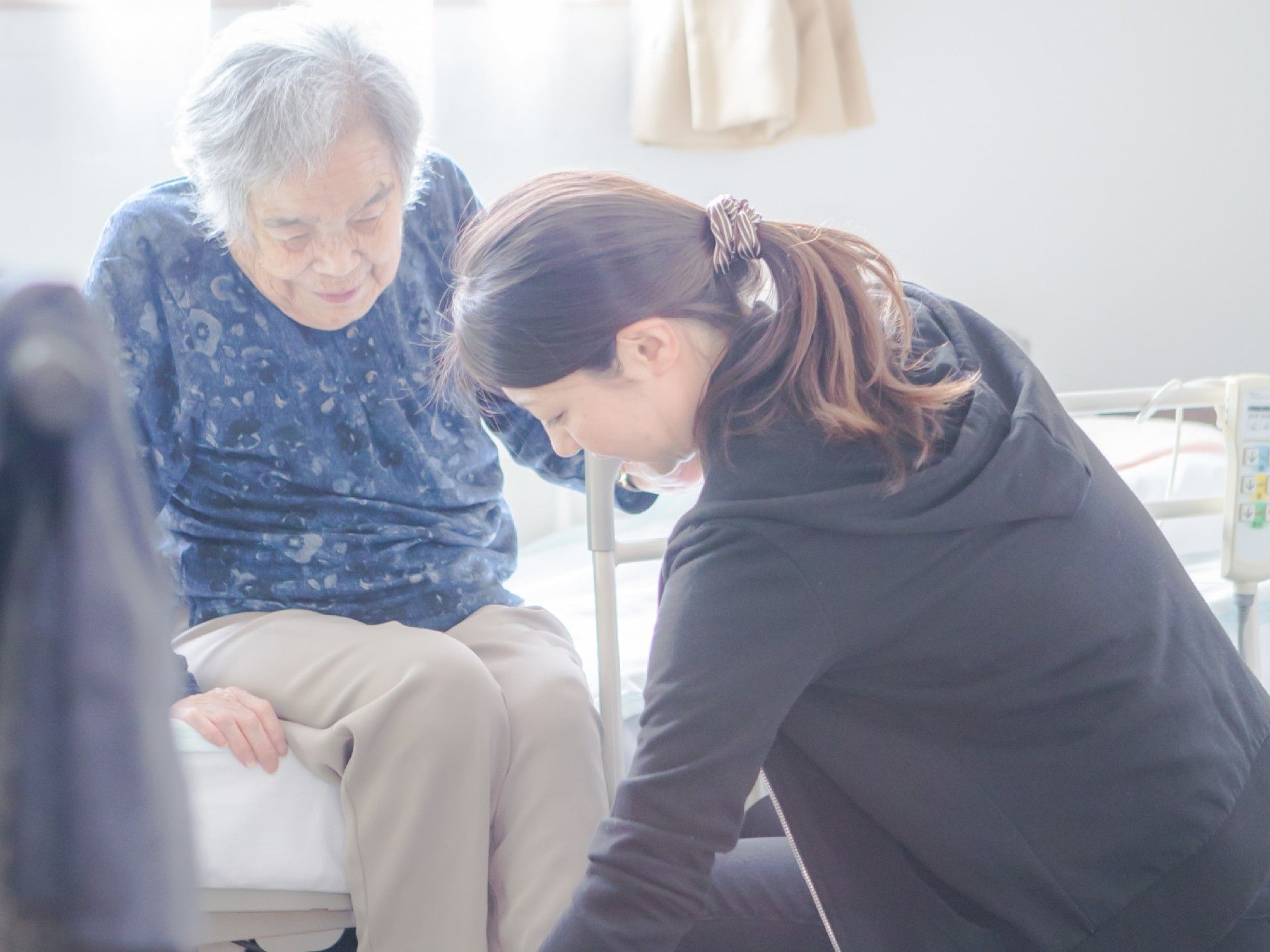 介護ベッドに座る高齢者の動作をサポートする、施設の女性スタッフ