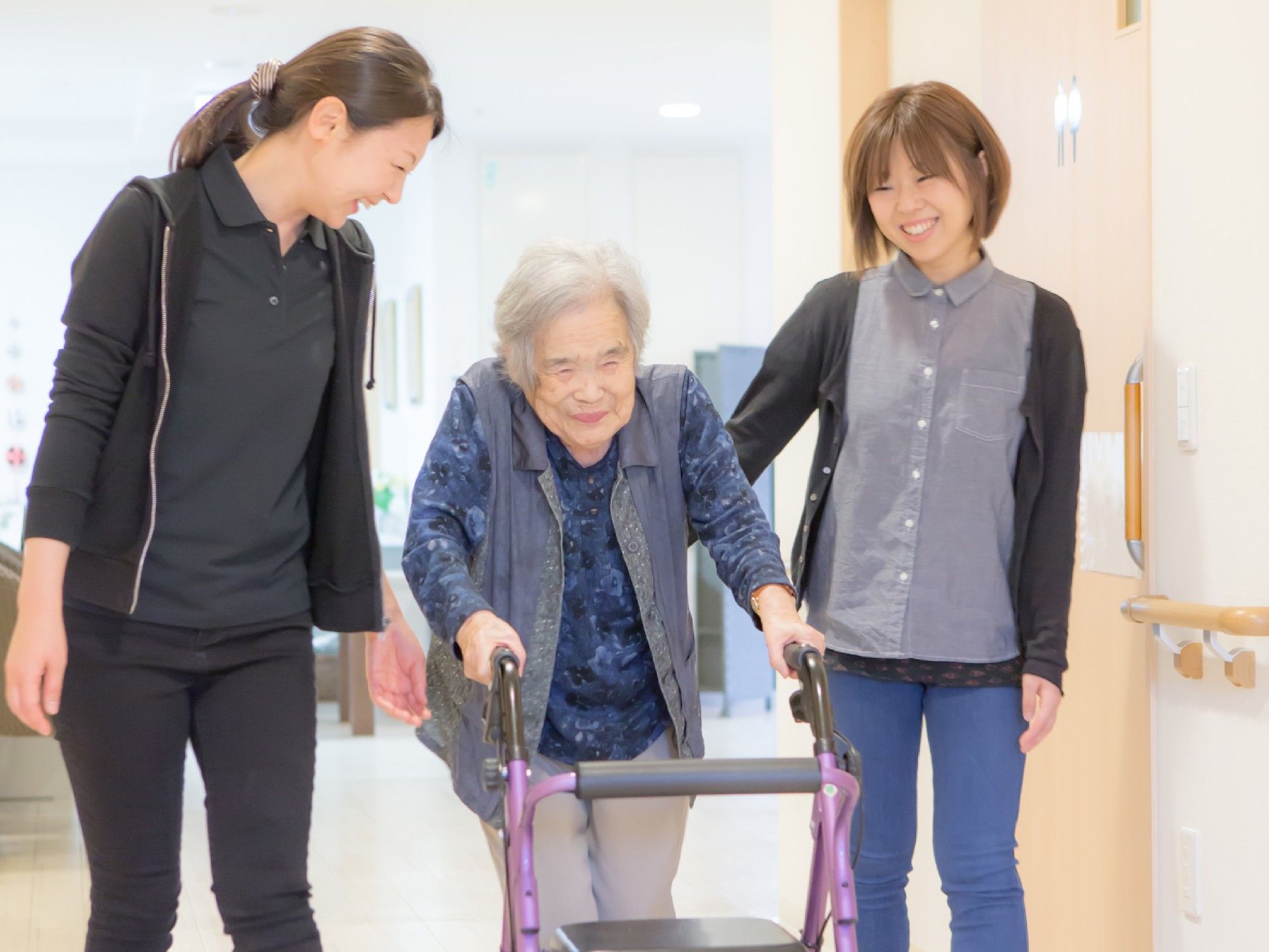 歩行器を押して歩く高齢女性を両側から見守る二人のスタッフ