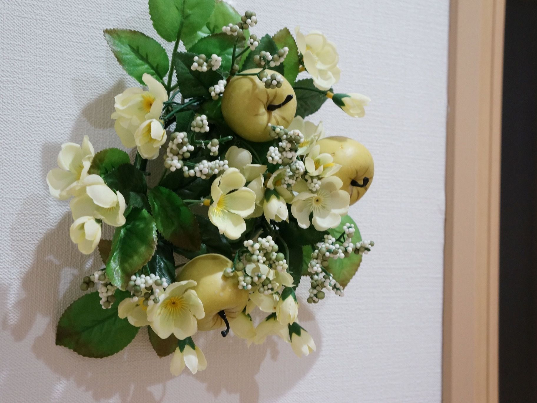 白色の壁に、リースの飾りが付いている。グリーンの葉っぱをベースにし、白い花、白い実、白っぽいリンゴがいくつか含まれている。