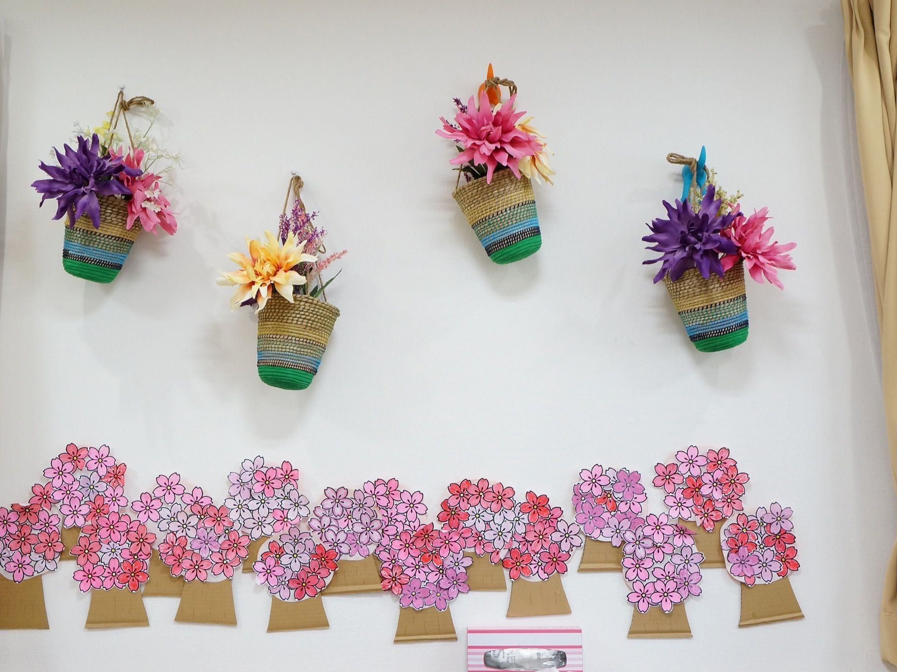 壁に、カゴに入った造花がいくつか掛かっている。その下には、桜の花の色を塗った作品が、たくさん貼られている。