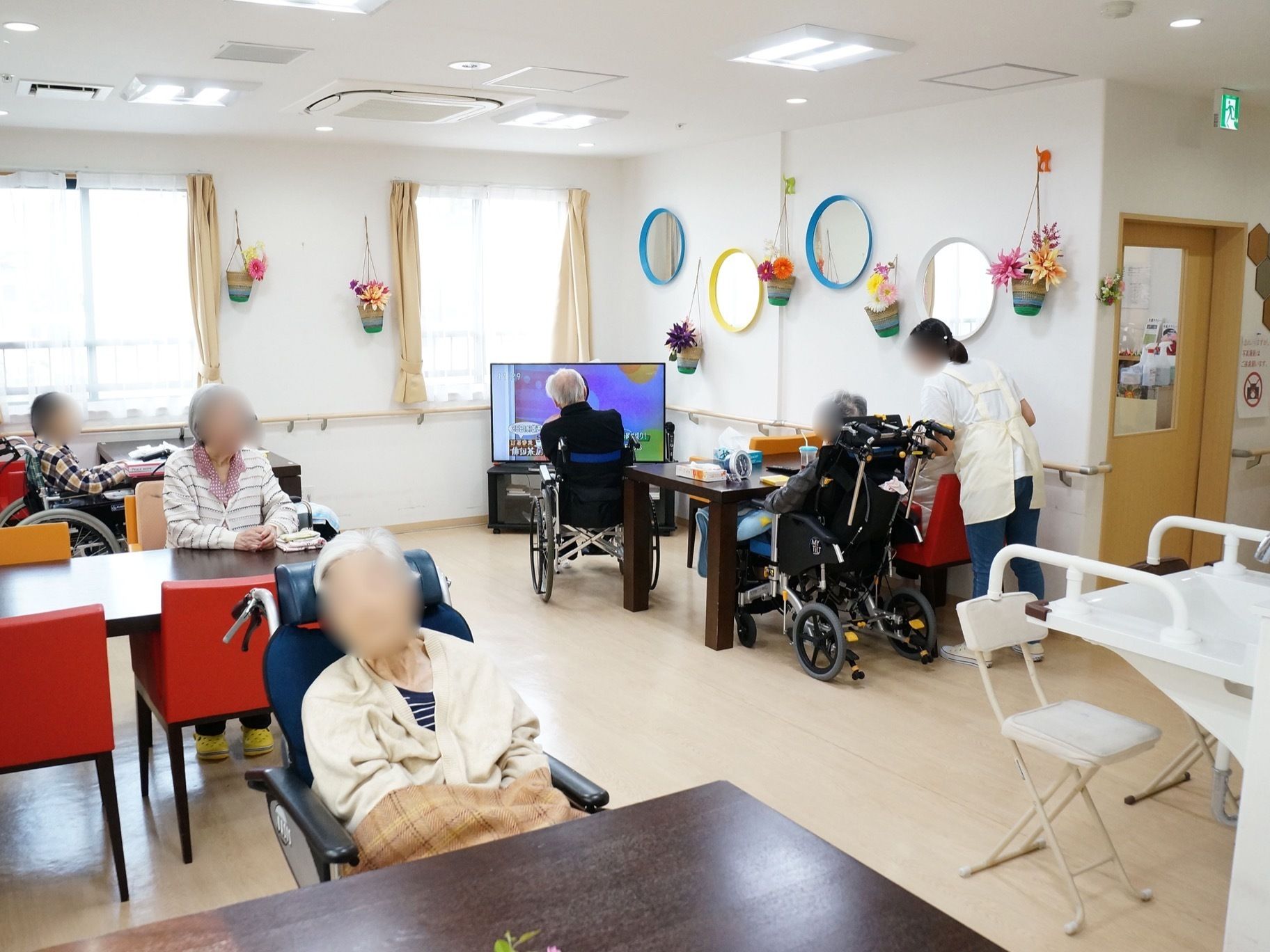 窓の多い明るい部屋に、テーブルや椅子が置かれている。部屋の角にはテレビが備わっている。手前には、洗面台が設置されている。