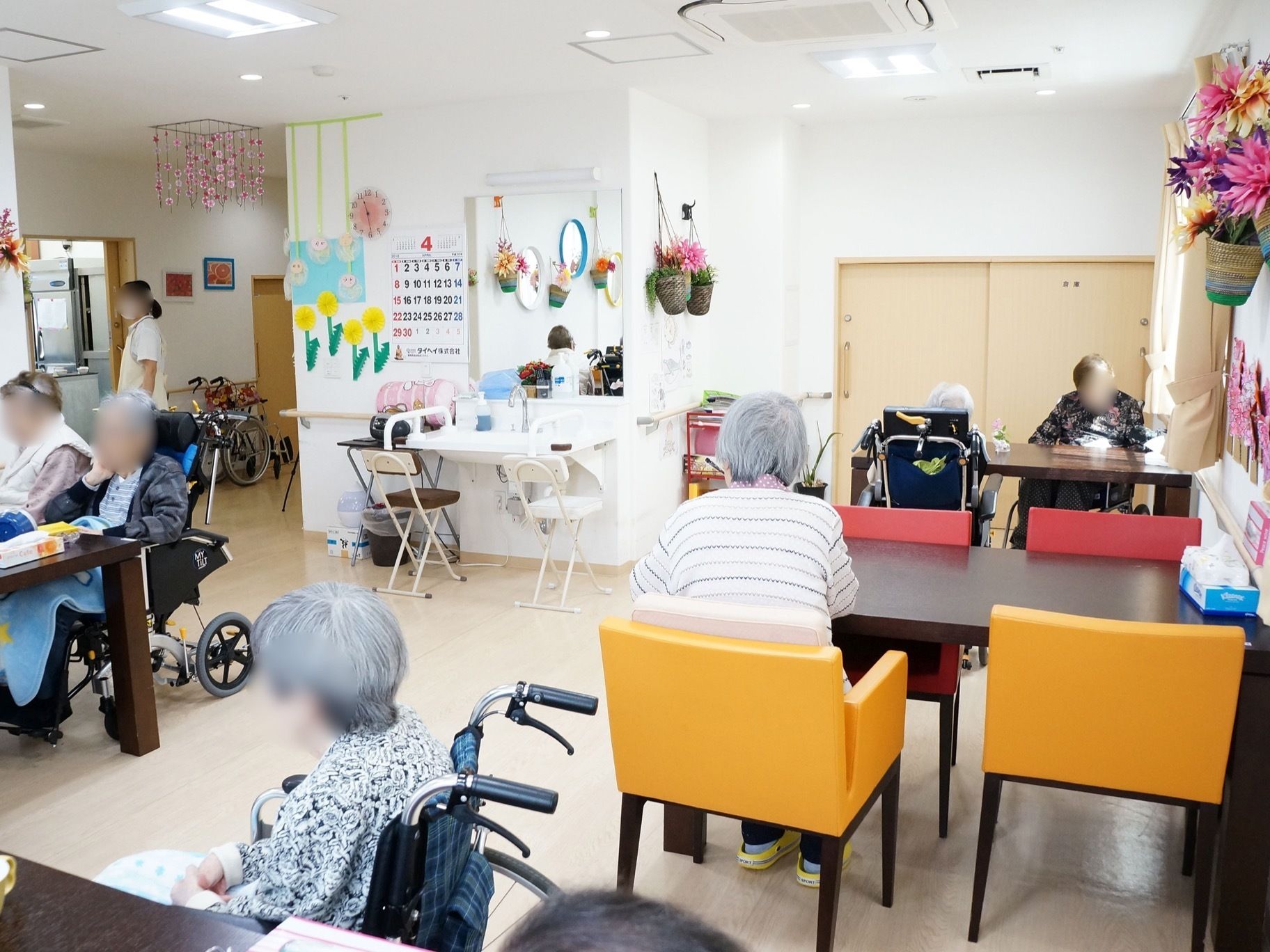 食堂の壁にはたくさんの飾り付けがしてある。入居者様がテーブルに付いている。奥の壁に、洗面台やカレンダーが設置されている。