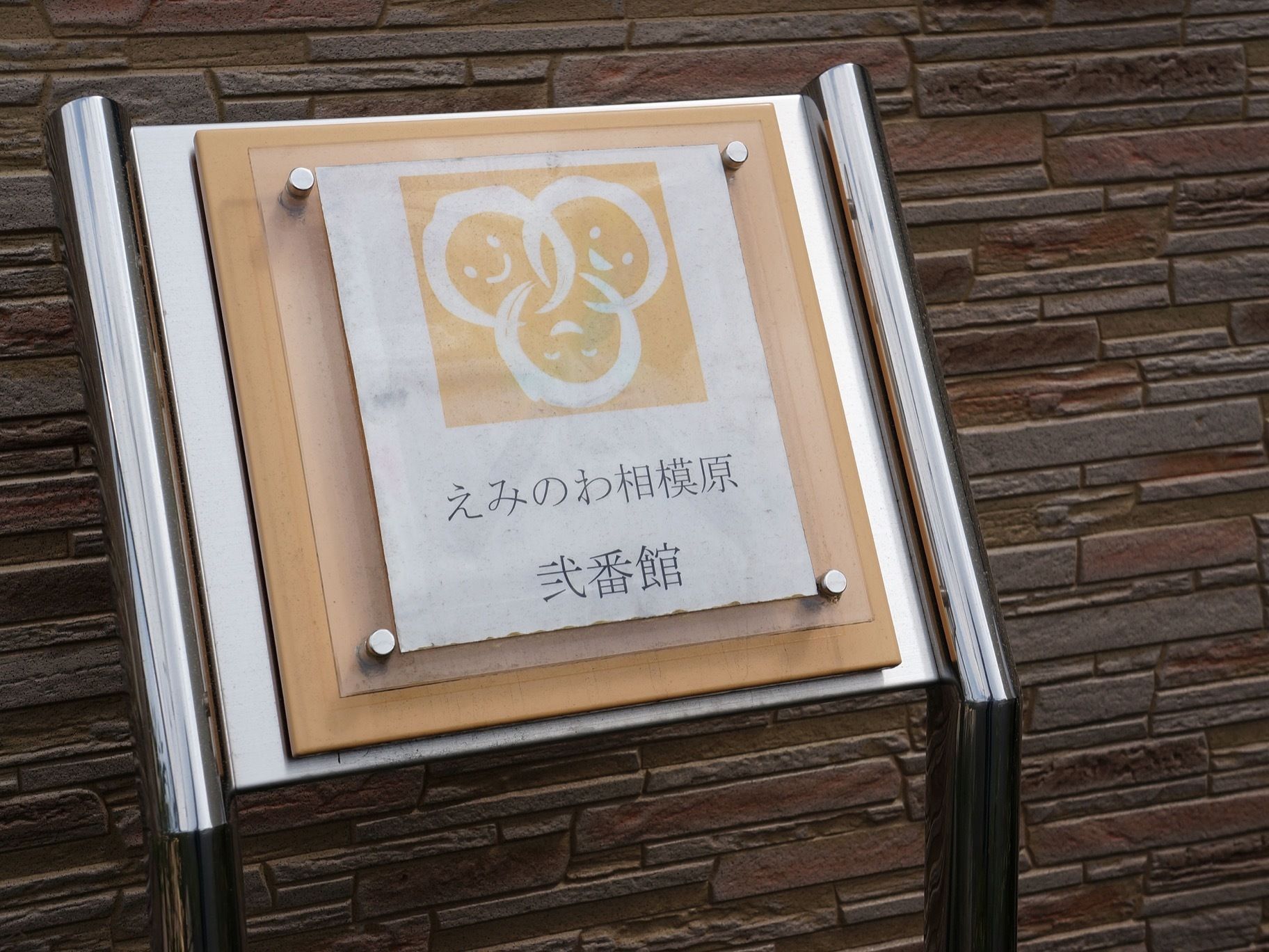 外壁の脇に、シルバーのポールで立っている看板がある。施設名とトレードマークが書かれており、バックは淡いオレンジ色である。