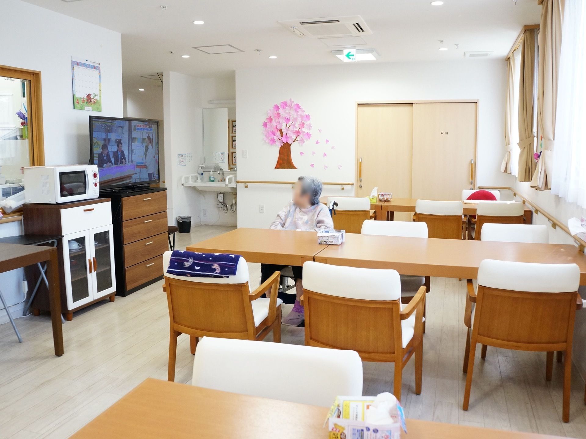 長方形の机が3列に並べて配置されている。各両端には椅子が置かれている。左の壁際にはテレビや電子レンジなどが設置されている。