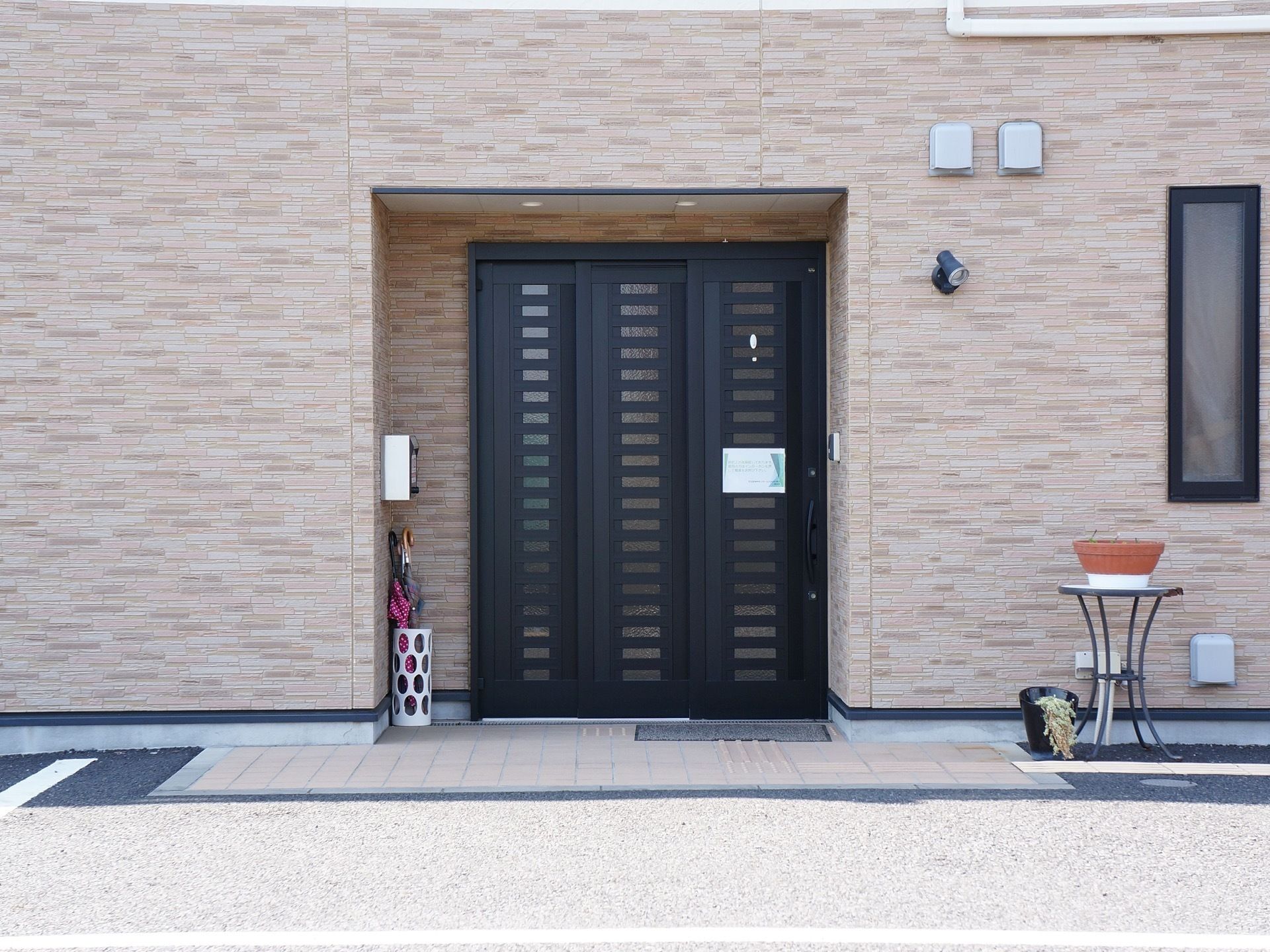 施設の外壁はレンガ調のようにみえる。壁のくぼんだ個所に黒いドアが設置されている。ドアの近くに傘立てがあるのが見える。
