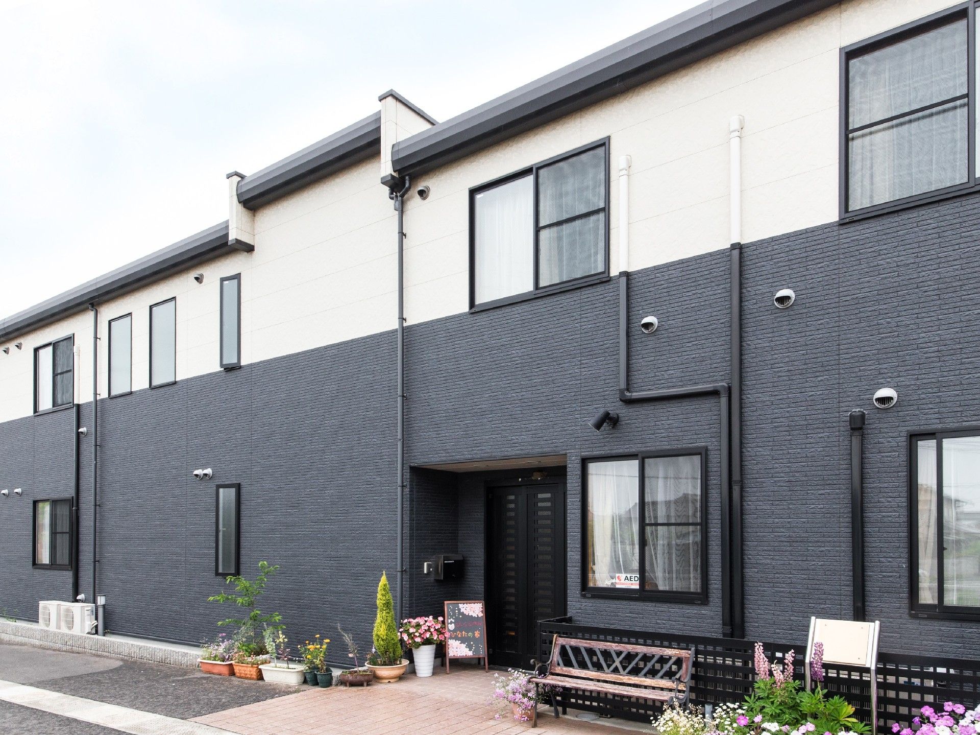 ２階建てのグループホームである。玄関横には、ウェルカムボードや綺麗な花が植えられた鉢植えが置かれている。
