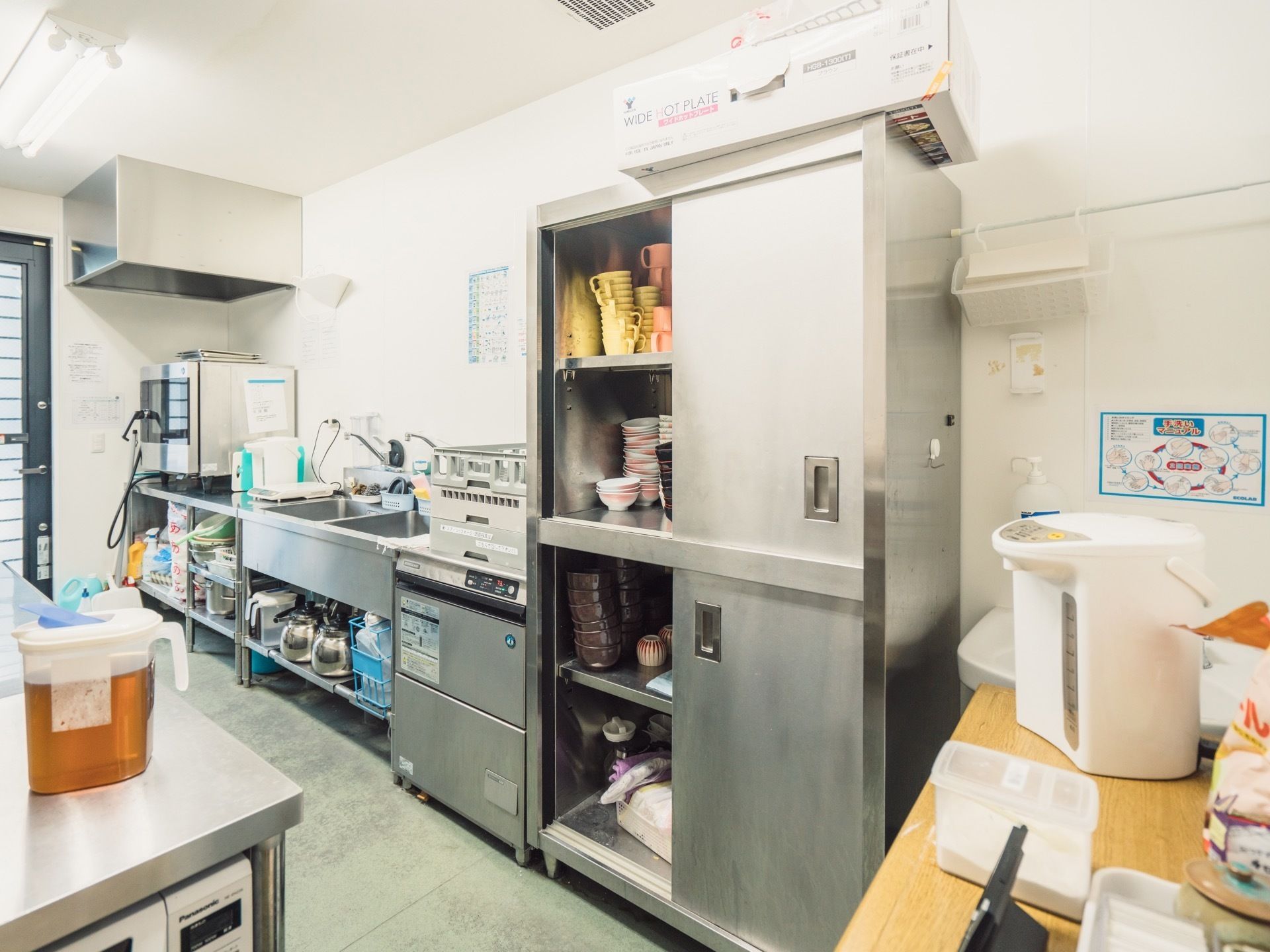 銀色の棚には食器やコップなどが入れられている。右にある木目調のテーブルには、電気ポットや小物が置かれている。