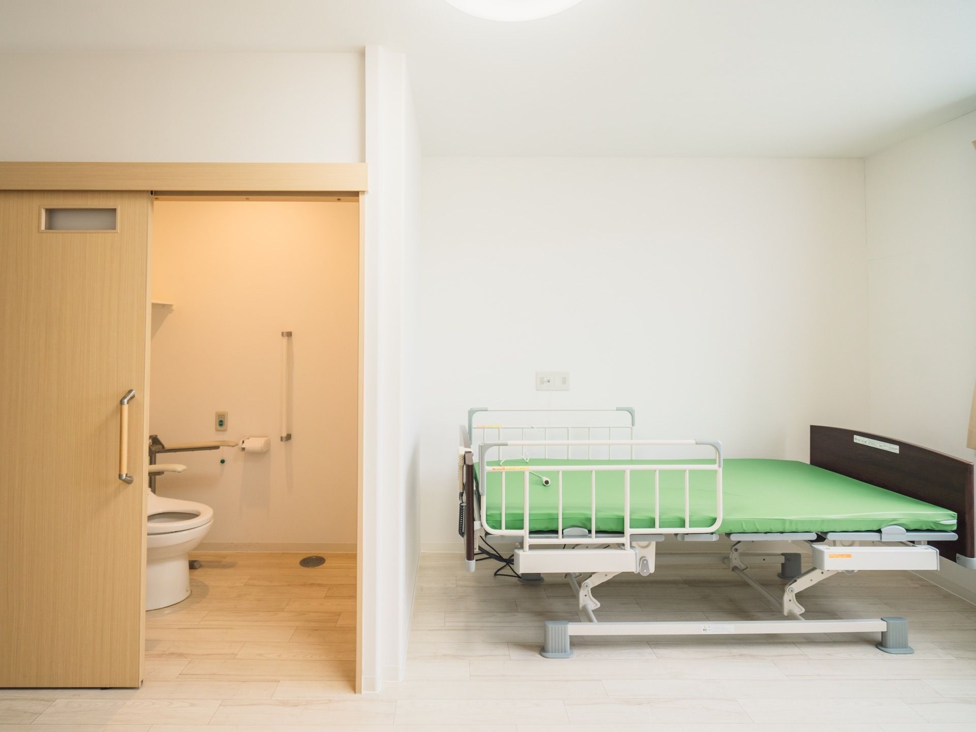 白を基調とした居室には介護ベッドが置かれている。隣にはトイレがあり、ドアは木目調の引き戸になっている。