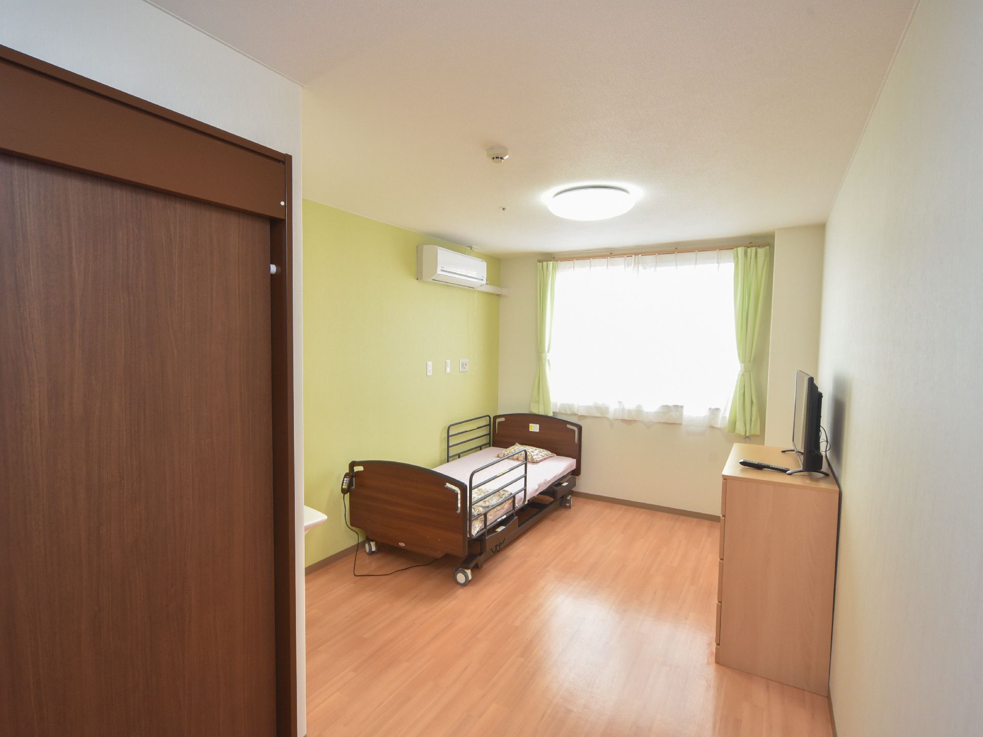部屋には腰高窓があり、明るい光が差し込んでいる。左側の壁とカーテンは、淡い緑色で揃えてある。電動式のベッドも置いてある。