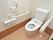 白で統一されたデザインのトイレは、フタが無いタイプとなっている。壁には大きな手すりがあり、オレンジ色の緊急ボタンもある。