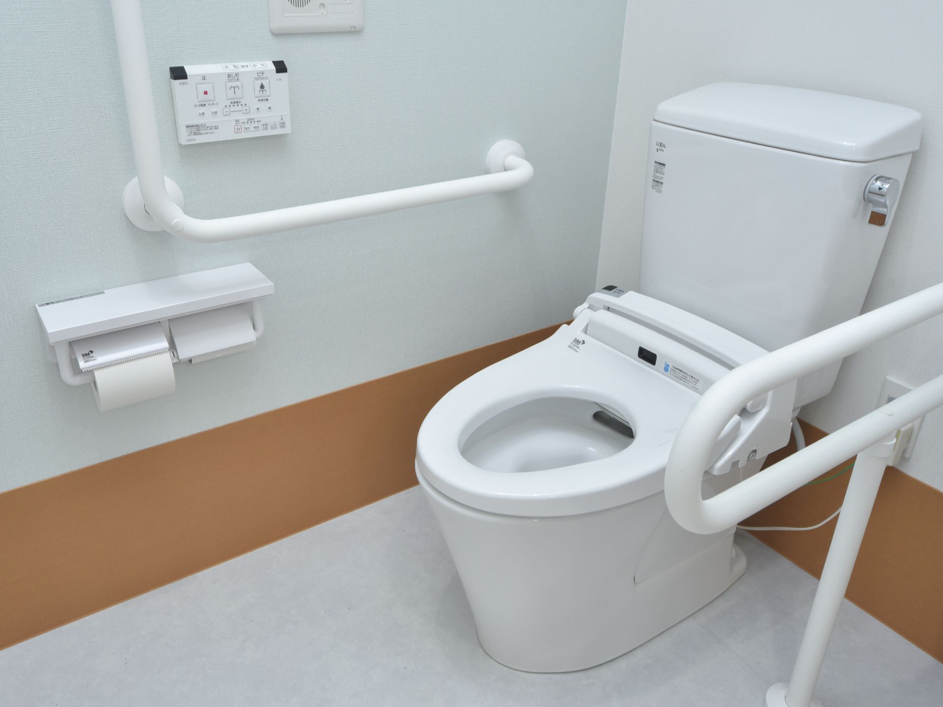 フタのないタイプの温水洗浄便器が設置されている。白い壁には、L字型の頑丈な手すりやトイレットペーパーが2個付いている。