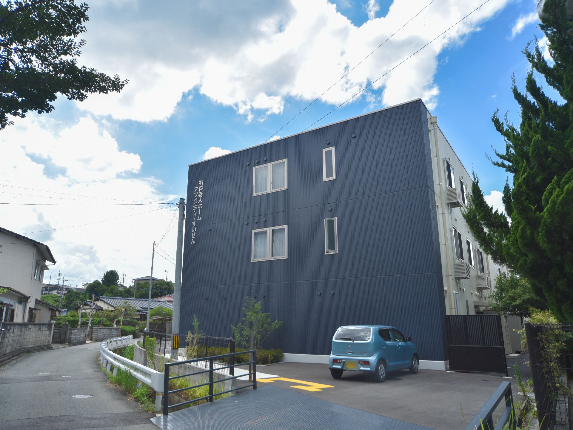 ブルー系の落ち着いた色合いの壁には、白い文字で施設名が書かれている。壁の前には、水色の軽自動車が置かれている。