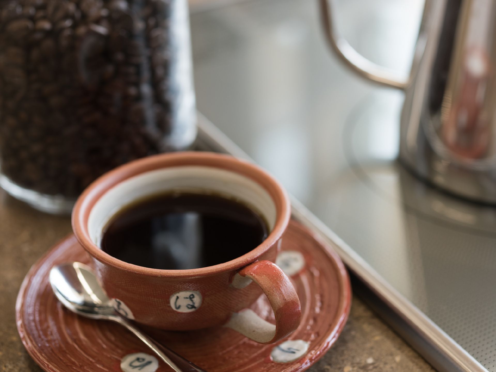 手作り風の陶器のカップに入れたてのコーヒーが注がれている。横にはコーヒー豆も置いてある。IHヒーターではコーヒー用のポットがある。