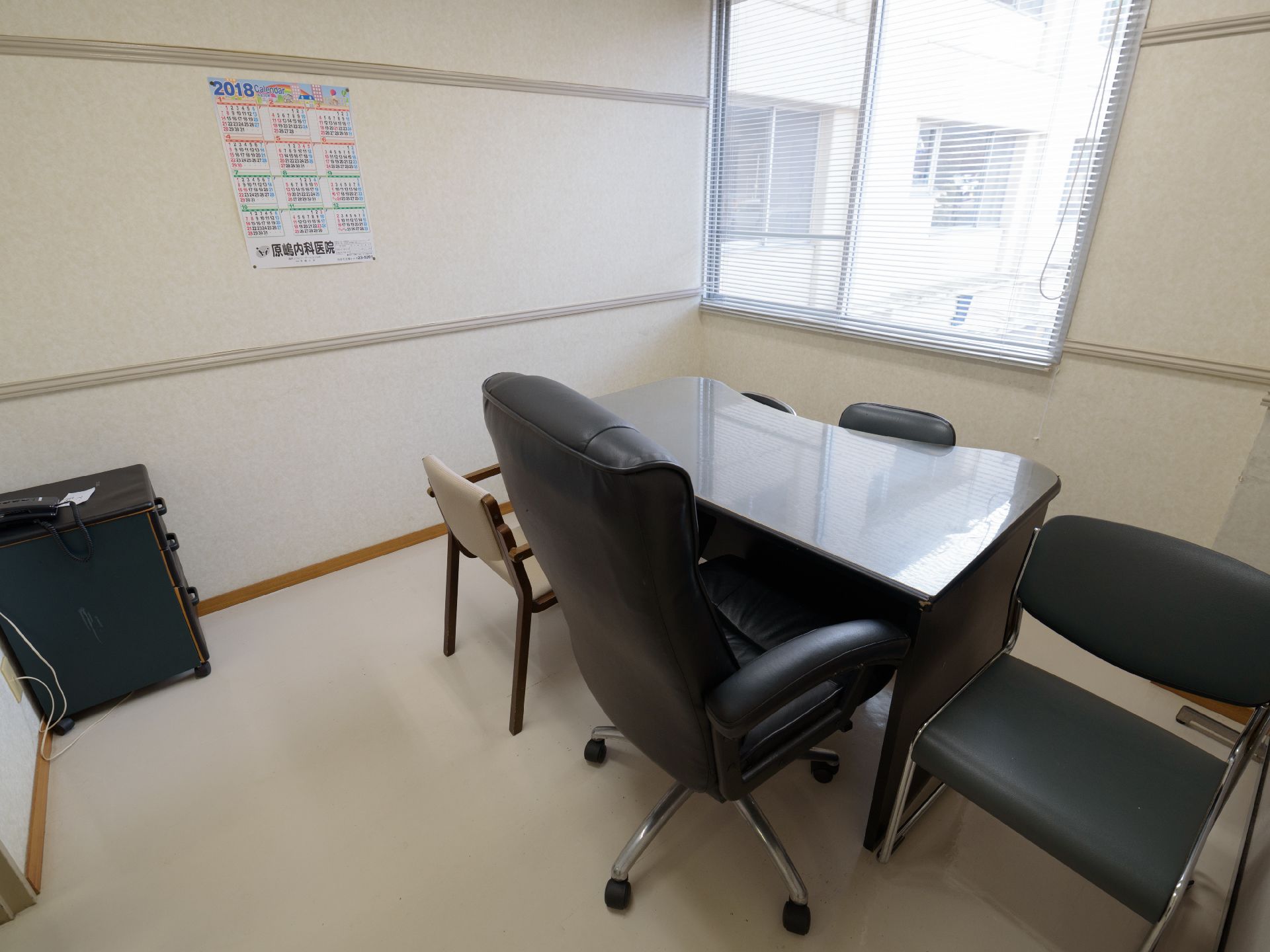 室内に黒いテーブルと椅子が置かれている。窓にはブラインドが掛けられており、壁にはカレンダーが貼られている。