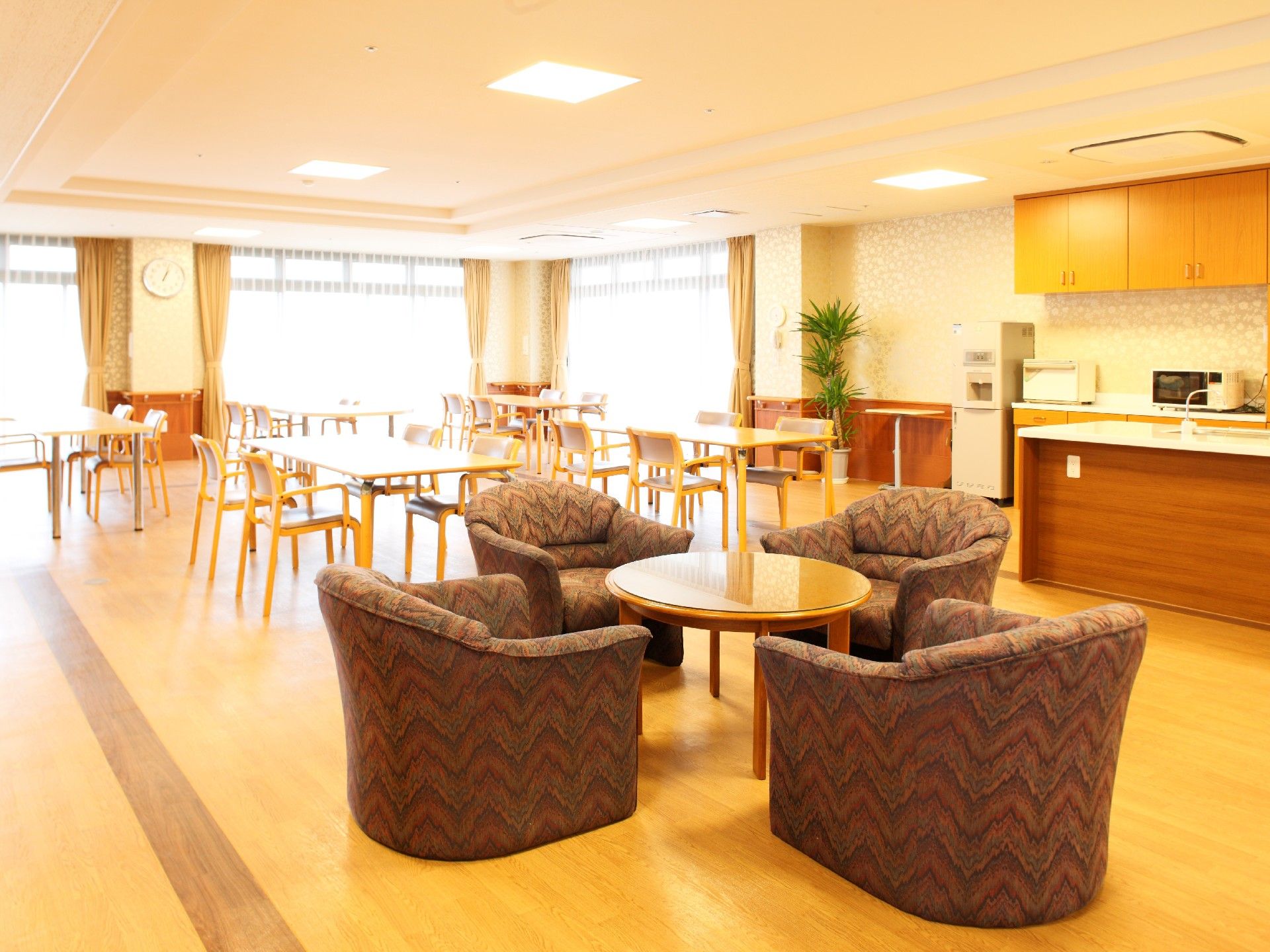 ２方向に窓がある明るい室内の窓側には、４人掛けのダイニングセットが並んでいる。手前には一人掛けソファ４脚とテーブルがあり、台所もある。