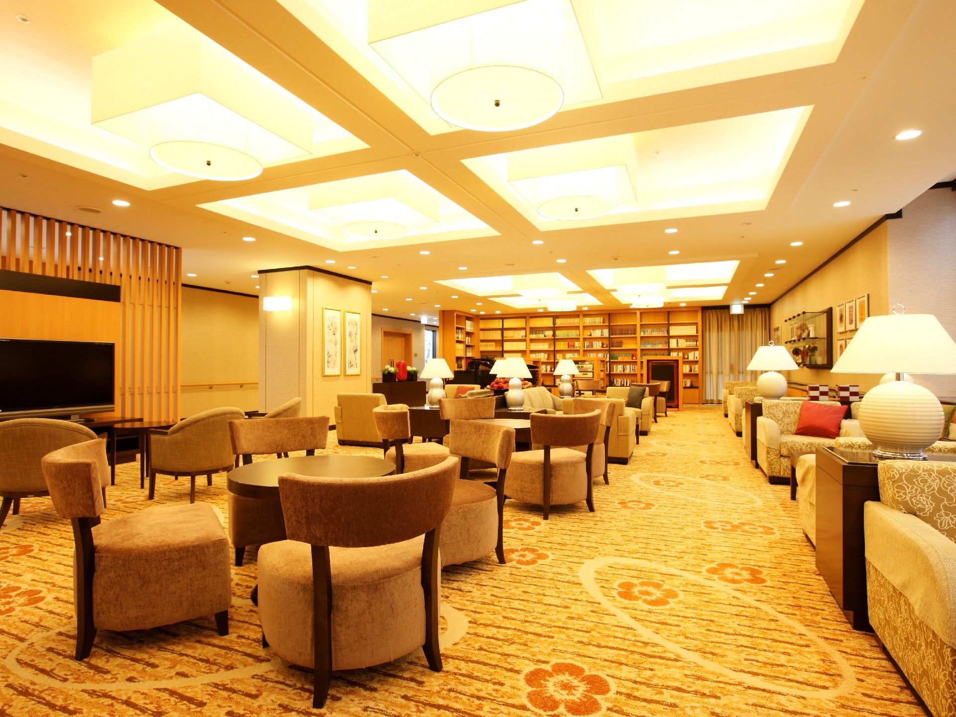 突き当りに大きな書棚がある。向かって右側はソファセットが並び、通路を空けて左側にはテレビと、丸テーブルを囲んで椅子４脚のセットが並んでいる。