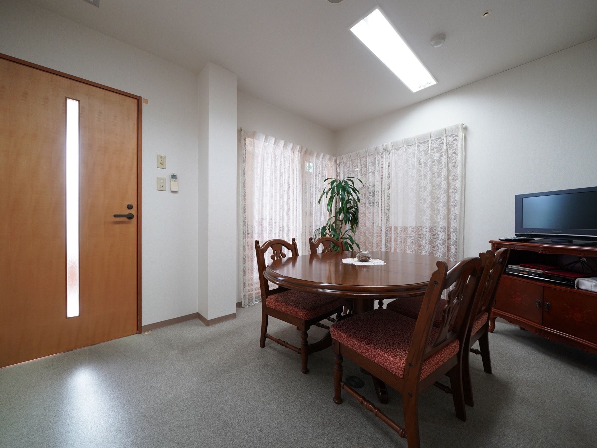 応接室は広々としていて、木目調のテーブルと椅子が４脚、テレビが設置されている。部屋の角に観葉植物が置かれている。