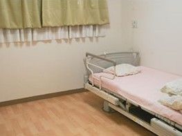 腰高窓があり、白いレースのカーテンと緑のカーテンが掛かっている。壁際には介護ベッドがあり、窓際には枕が置かれている。