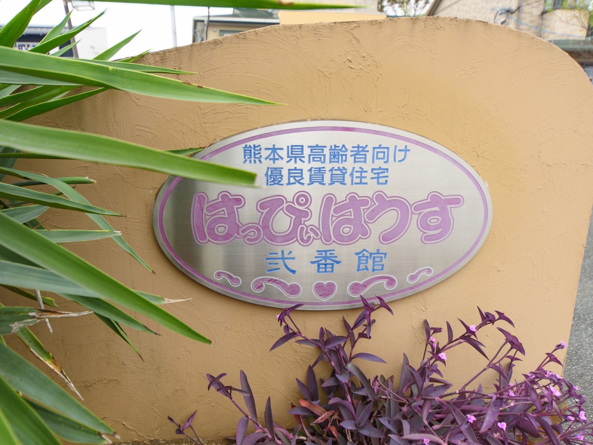 施設の入口にある塀には、施設名が書かれたプレートが取り付けられている。熊本県に位置し、高齢者向け住宅施設である。