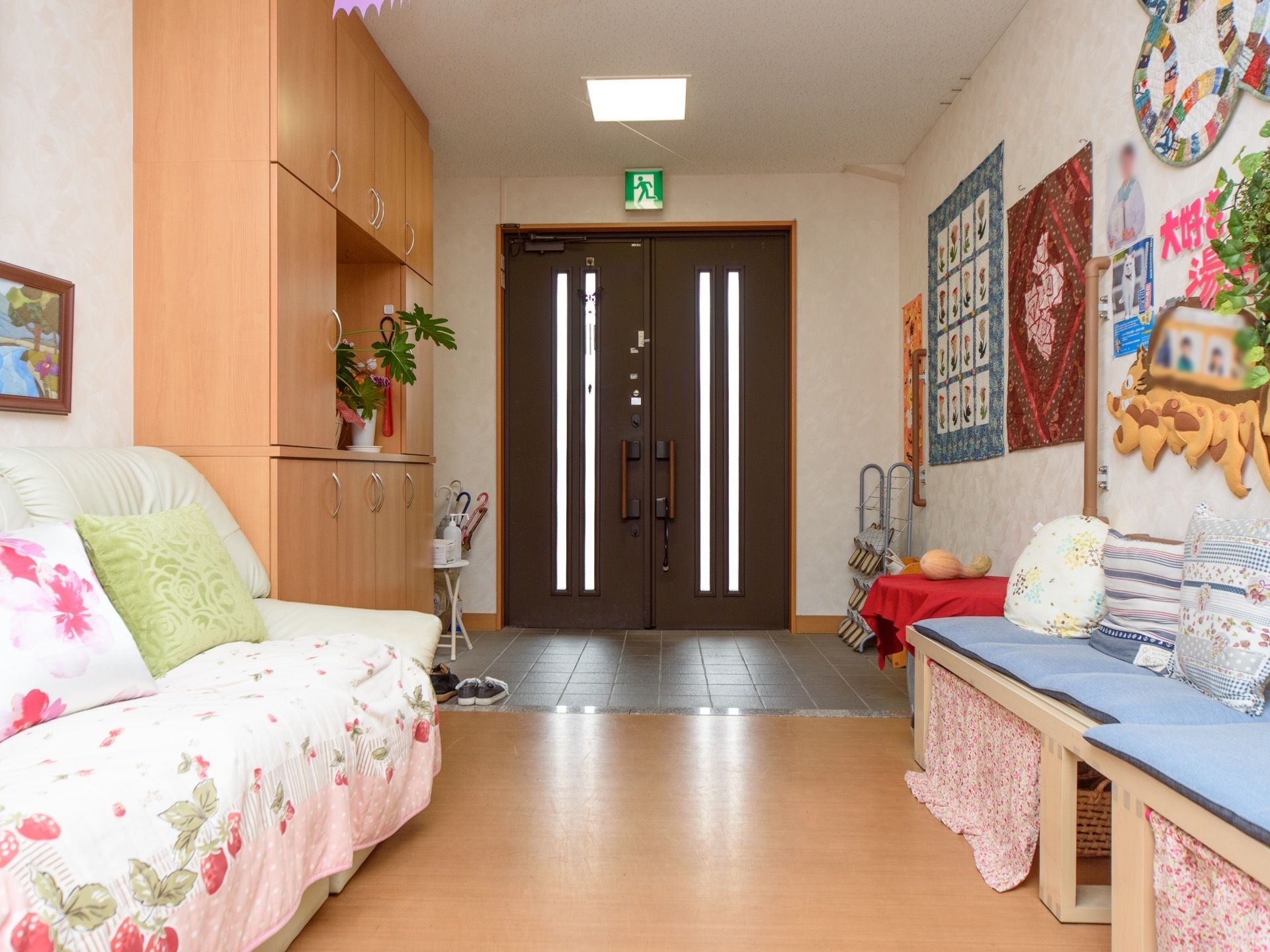 玄関は間口の広いドアで段差がない。備え付けの大きな下駄箱がある。椅子やソファが置かれ、壁にはキルトが飾られている。