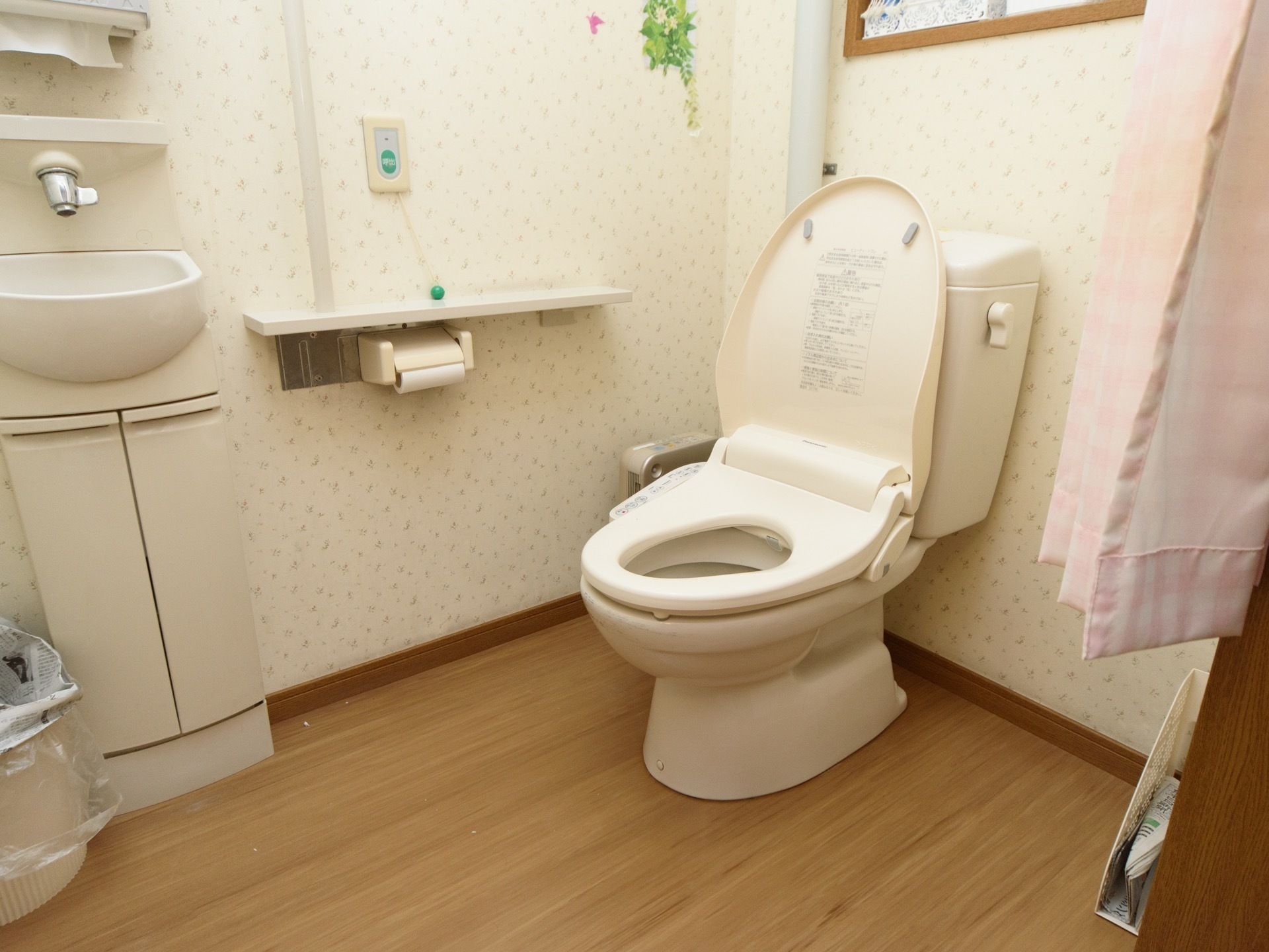 入り口にピンク色のカーテンが掛けられたトイレがある。壁紙は花柄で明るく、壁には手すりとナースコールが設置されている。