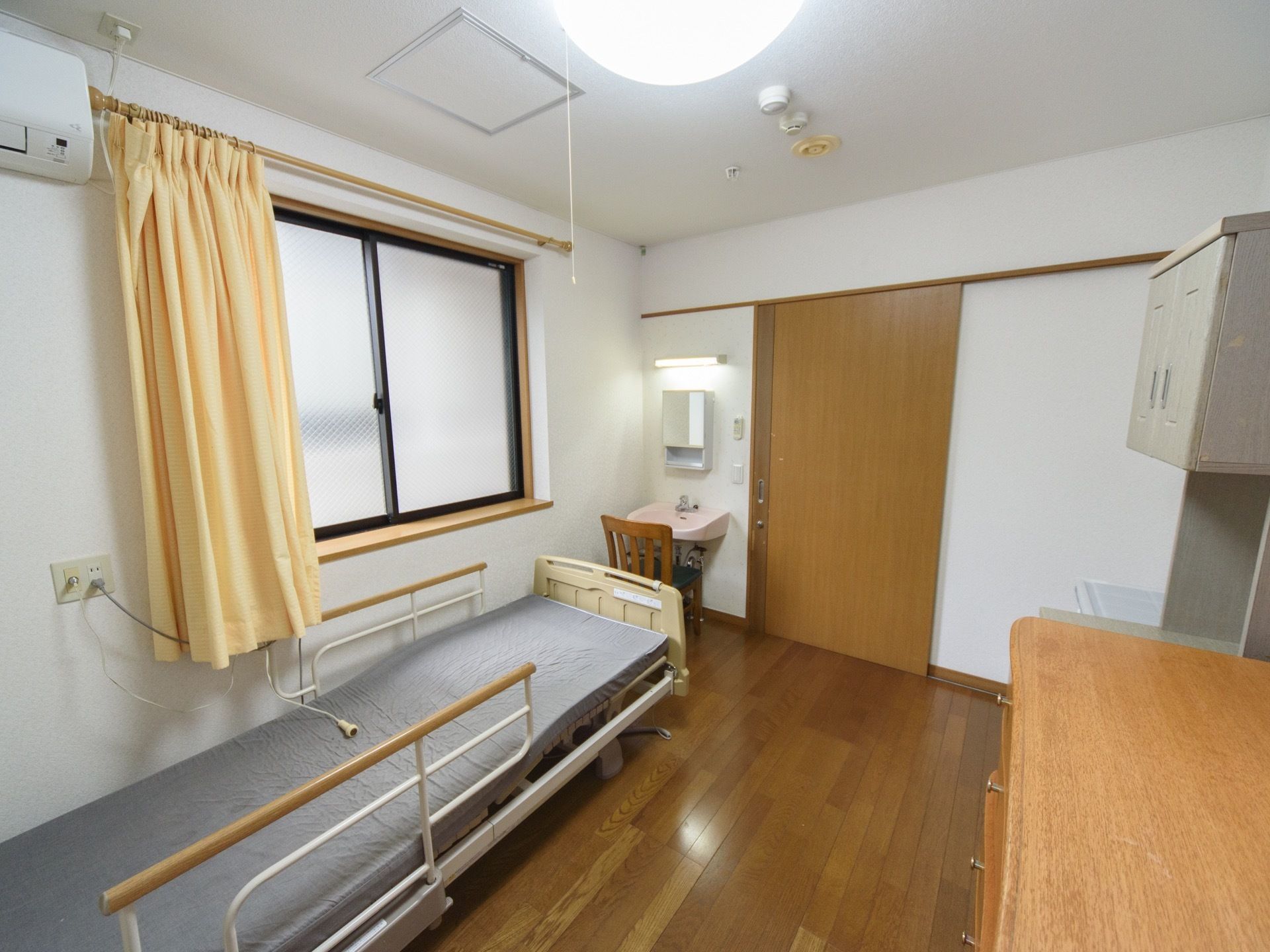 フローリングの居室に電動ベッドが置かれている。黄色いカーテンがかかった窓があり、その横にはエアコンを設置。