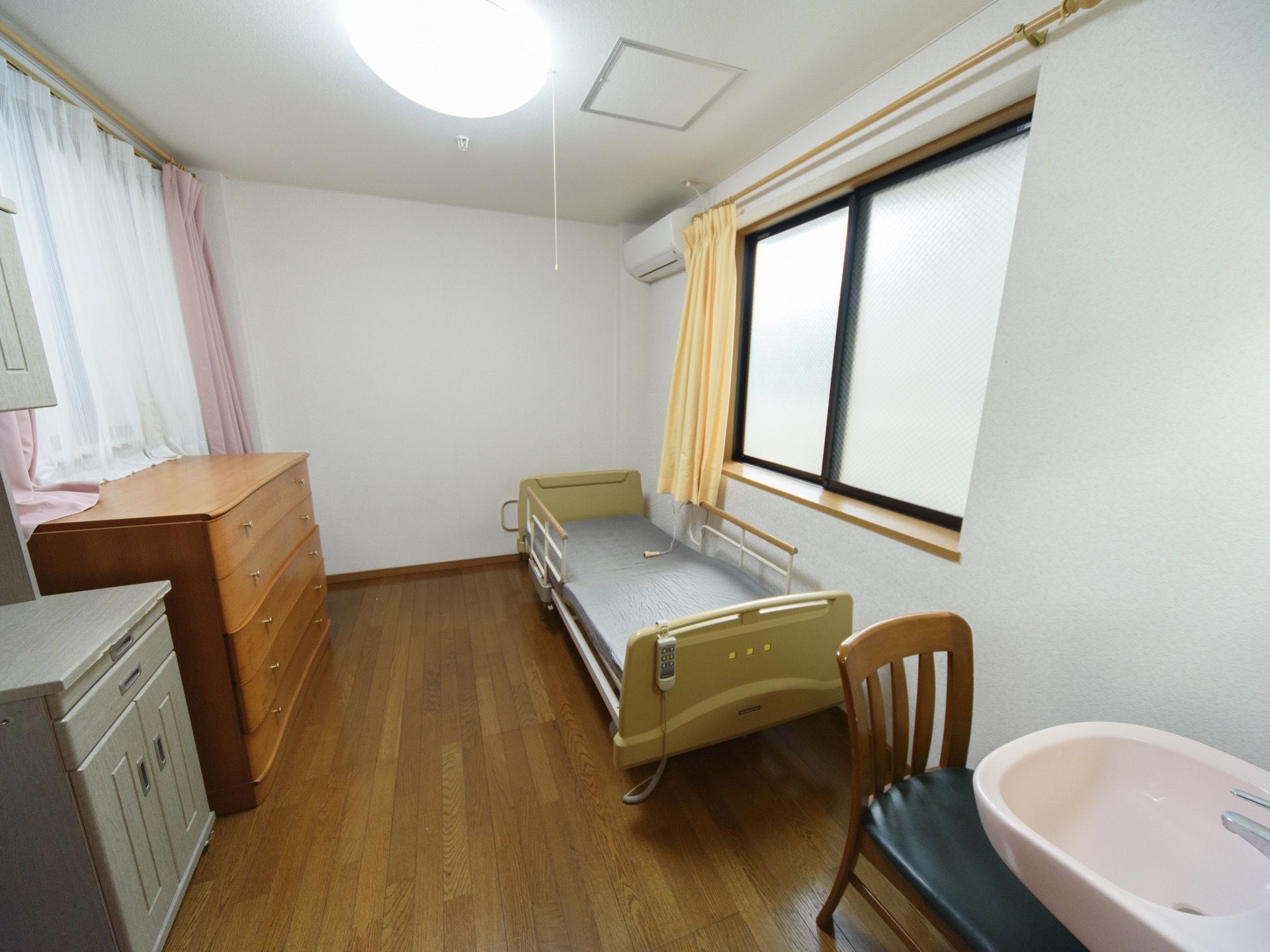 フローリングの居室に、木製のチェスト、電動ベッドが置かれている。部屋の両側に窓があり、それぞれ黄色とピンクのカーテンが掛かっている。
