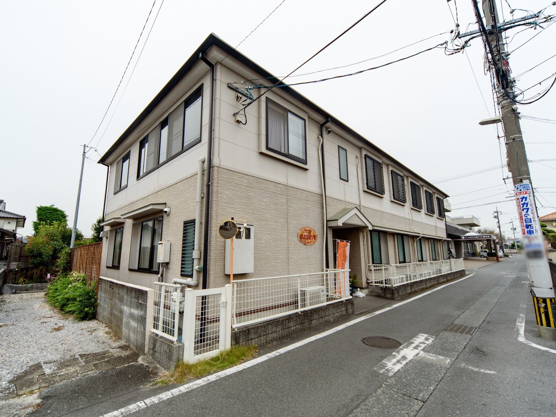 白とグレーを基調とした２階建てのアパートが立っている。車道から玄関までは緩やかなスロープになっている。