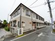 白とグレーを基調とした２階建てのアパートが立っている。車道から玄関までは緩やかなスロープになっている。