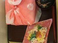 菱形の入れ物に入ったちらし寿司がトレーに乗っている。一緒にお吸い物の椀と、桃色の風呂敷包みが置かれている。