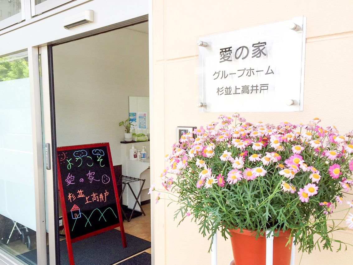 大きな花の鉢植えが置かれたエントランス。「ようこそ愛の家杉並上高井戸」と書かれたブラックボードも置かれている