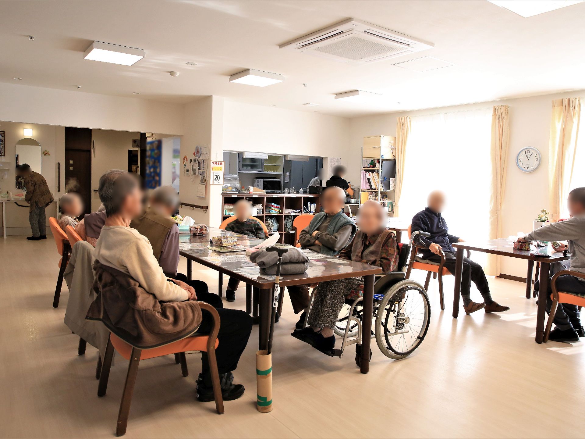 ダークブラウンのテーブルセットに入居者様が座って談笑している。奥にはキッチンがあり、スタッフが作業をしている。