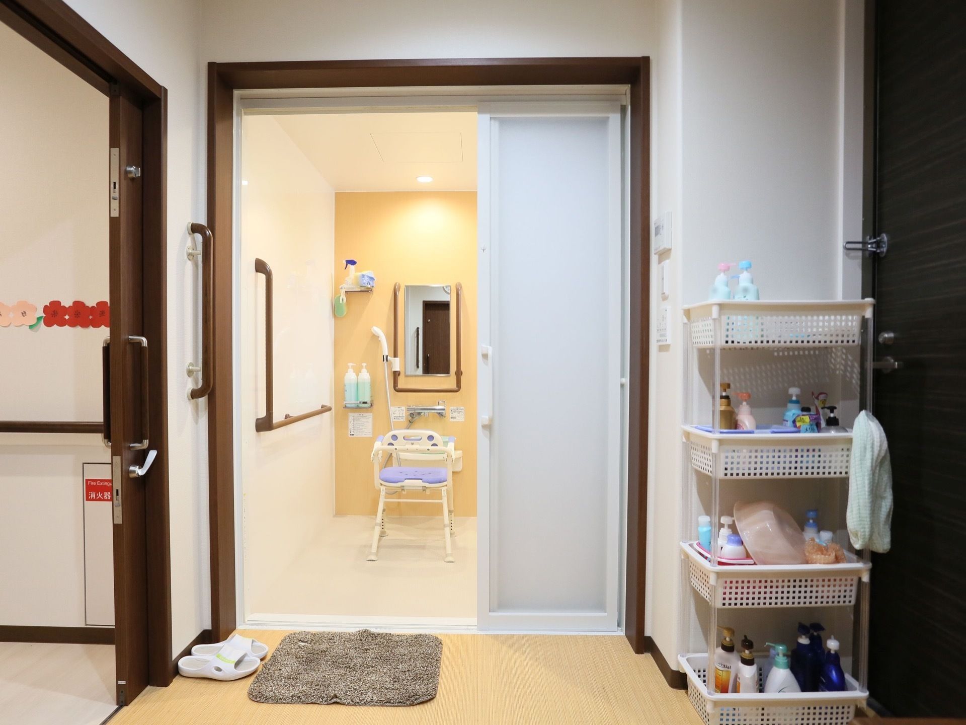 右端に４段の白いワゴンがあり、さまざまな種類のシャンプーやリンス、洗面器などが置いてある。床には、マットとスリッパもある。