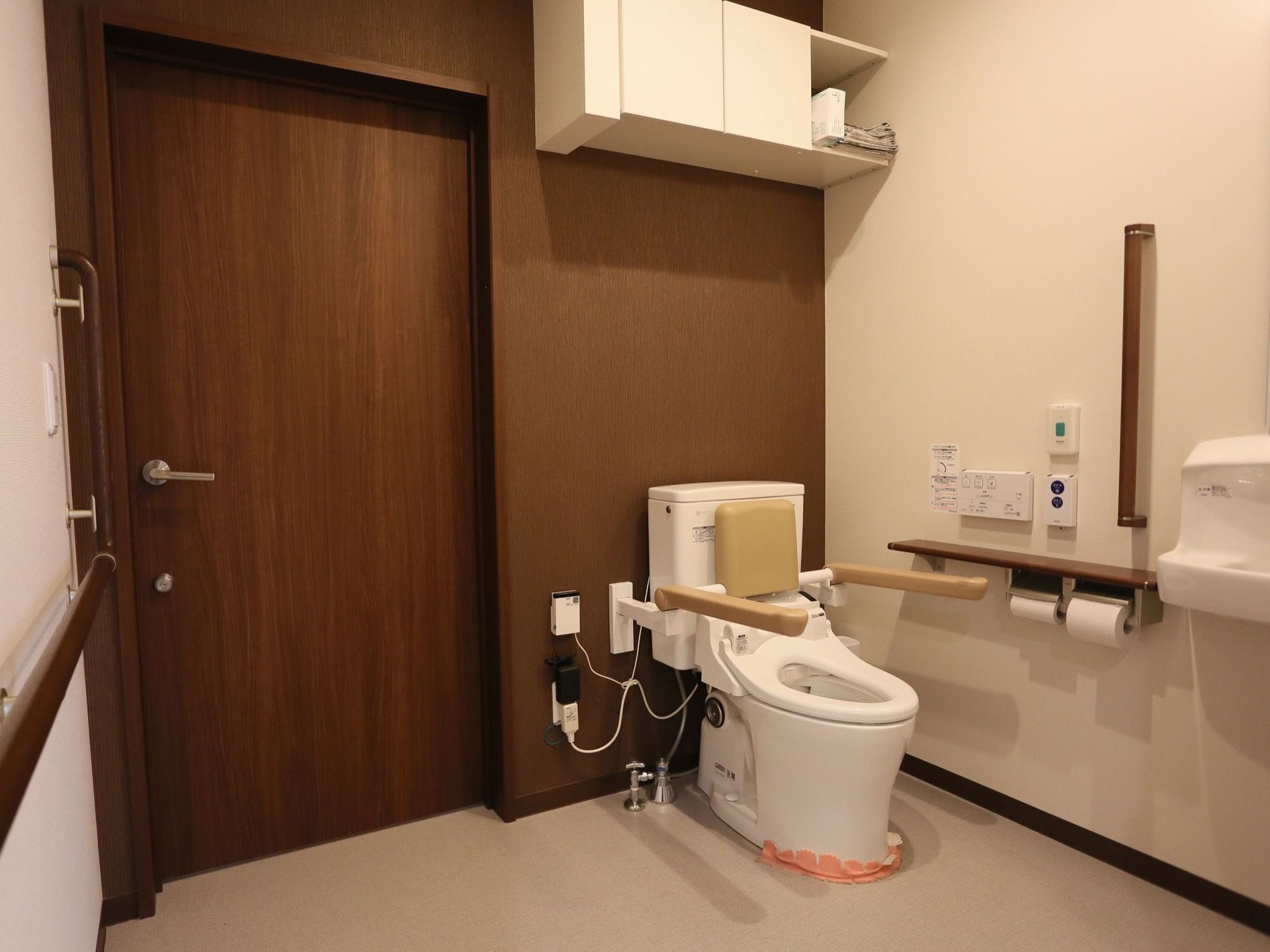 広い空間が確保されているトイレである。ベージュ色の手すりと背もたれが設置されている。上の方には、収納棚もある。