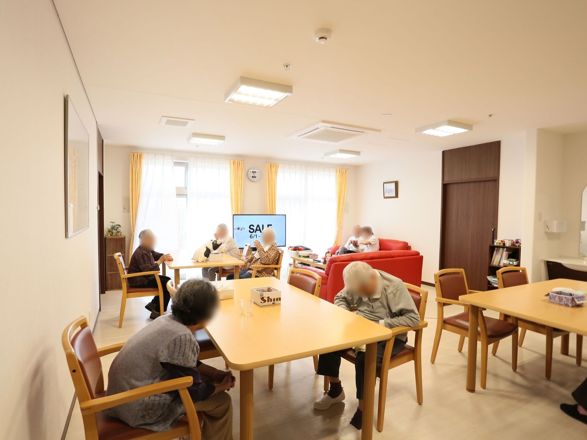 食堂の奥には大型の窓があり、明るい光が差し込んでいる。数名の入居者様がテーブルの周りに座って、談笑したりテレビを見たりしている。