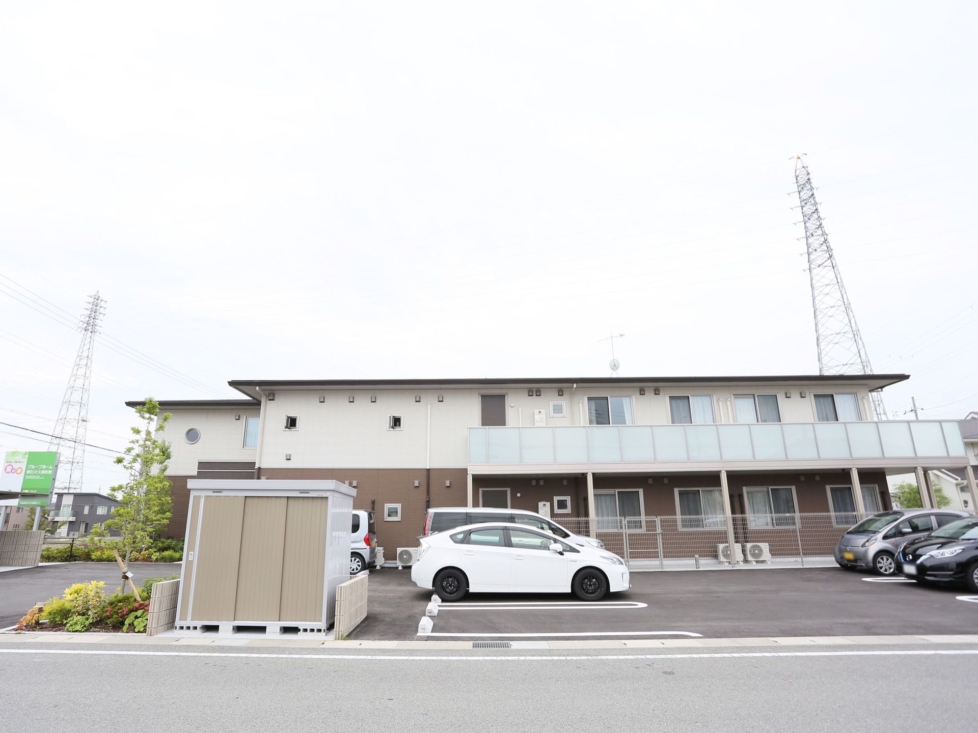 地上２階建ての施設の前方に、広々とした駐車場が設けられており、数台の車が置いてある。左側には、植込みがある。