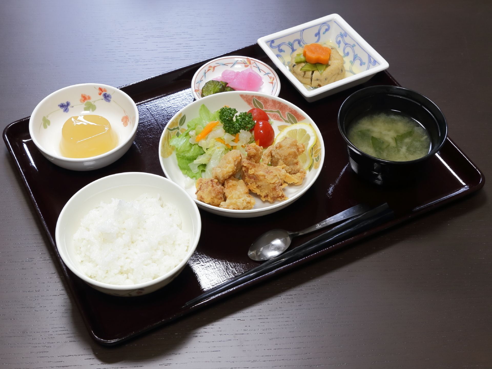 黒いお盆にご飯、みそ汁、メイン料理の鳥の唐揚げ」、がんもどきの小鉢、漬物、デザートが用意されている。